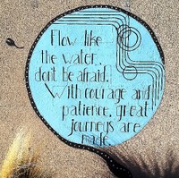 A blue circle painted on an adobe wall and adorned with a black outline and black, geometric line motifs along with the text "flow like the water. don't be afraid. with courage and patience, great journeys are made."