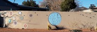A wide view of an adobe wall with various painted artworks, including birds on power lines, a blue circle with a poem written in black in the middle, and a tree with long branches that have colorful circle artworks on the ends of each.