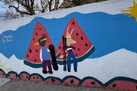 A wall mural painting of two, humanoid-style entities (one with a bird head, the other with a dog head and tail) standing in front of huge watermelon slices.