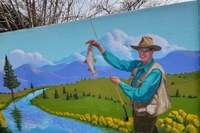 A wall mural painting of a person holding up a fish in one hand and holding a fishing pole the other. In the background is a nature scene with a river, grassy fields, and mountains in the background.