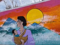 A wall mural painting of a mother holding an infant in the foreground and a sun setting over blue mountains in the background.