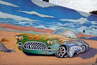 A painted wall mural of a green hot rod car driving along a desert with a big blue sky and white clouds in the background.