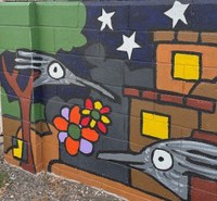 A wall mural painting of a simplified, stylized night time neighborhood with buildings, trees, stars in the sky, flowers, and two roadrunner heads poking out from behind a building and a tree.