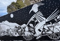 A stylized wall mural painting in black and white featuring a roadrunner riding a bike down a path at night with mountains and a starry sky and the moon in the background.