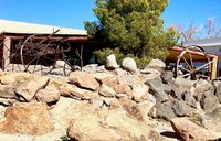 A pile of rocks and a tree in front of a house with a long, wooden covered porch.
