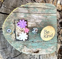 An old wood panel with severely faded teal paint and several aged embellishments like fake flowers and a "be kind" pin.