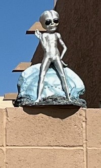 An alien sculpture standing in front of a small flying saucer.