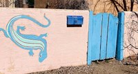 A painting of a teal lizard on a light tan adobe wall.