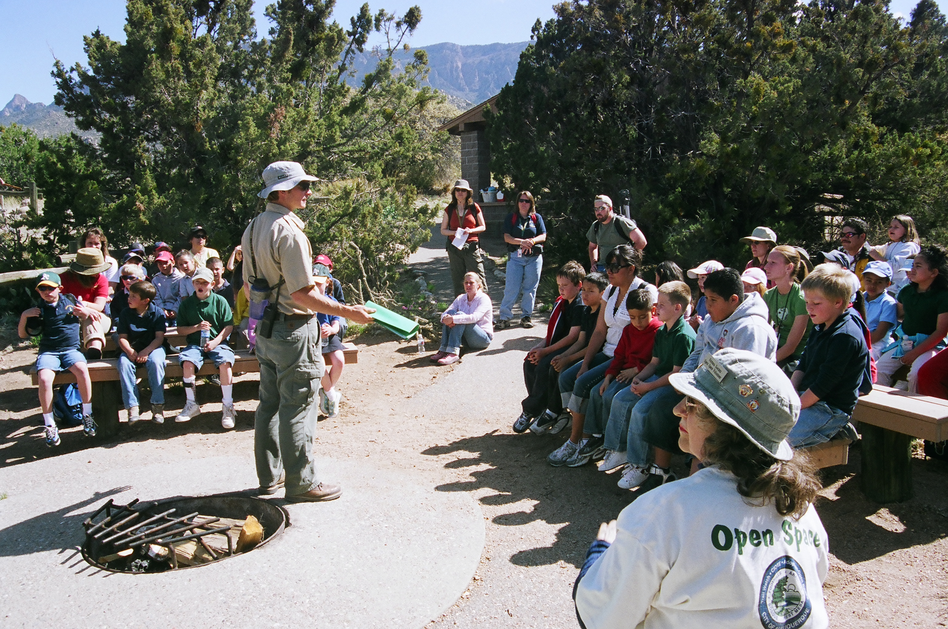 A group of kids and adults sitting on benches around a fire pit listening to an Open Space volunteer teach.