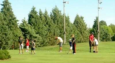 A group of children and adults playing golf on a green field surrounded by pine trees.