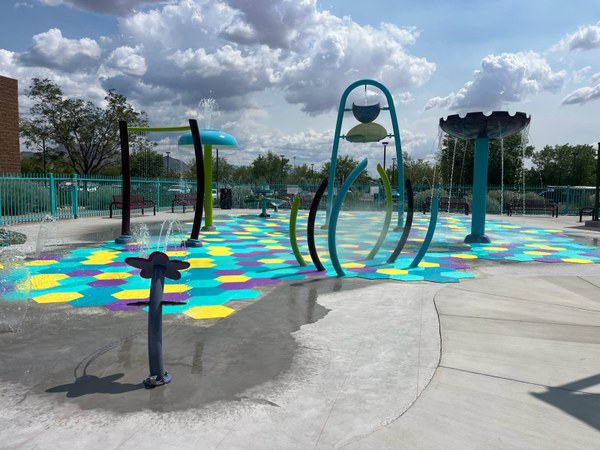 The Manzano Mesa Spray Pad, featuring a concrete area with multi-colored hexagonal tiles lining the floor, and multiple metal water spouts resembling flowers and mushrooms with water flowing down and out.