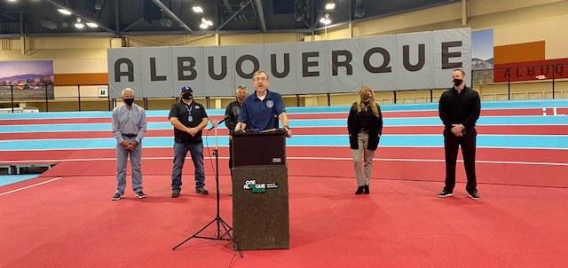 Director Simon standing at a podium in front of 4 other people and a banner that says Albuquerque.