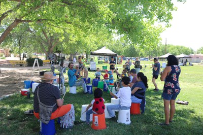 A group of youth and adults sit on buckets in a park banging on drums during a free lesson.