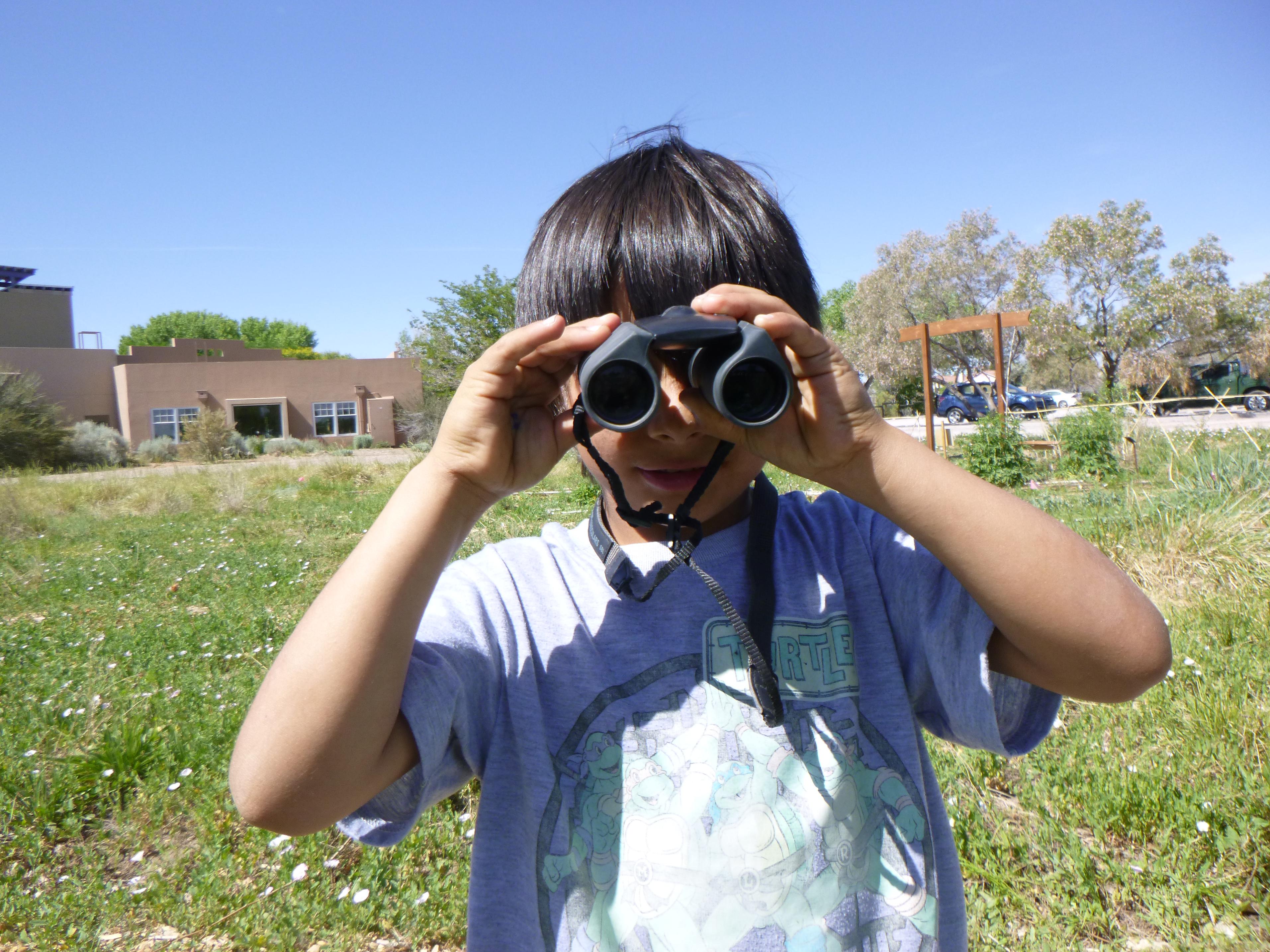 Image of a young student standing outside near the Open Space Visitor Center building. The student is holding up a set of binoculars and facing forward.