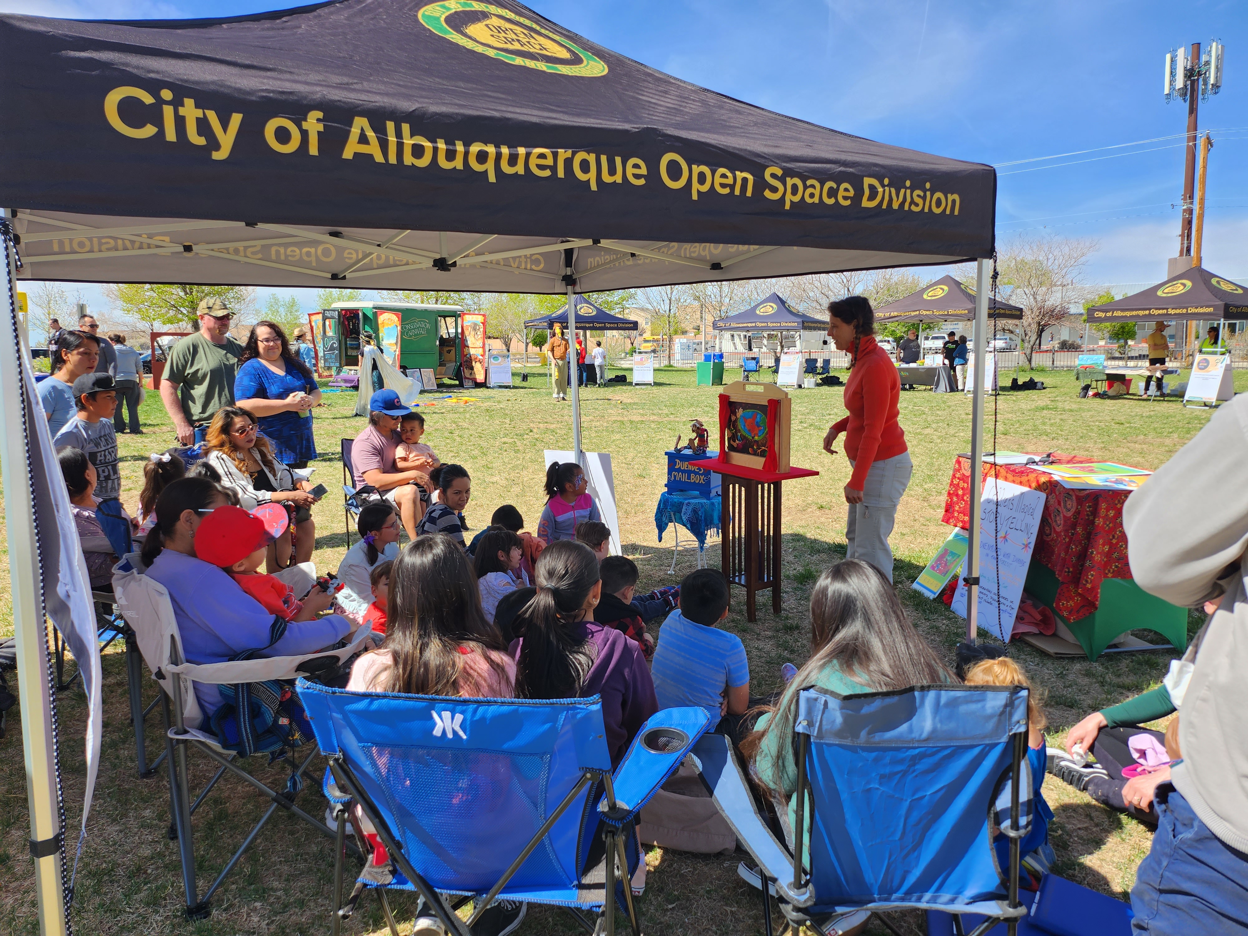 Open Space community event outdoors on grass. An audience sitting in lawn chairs under an Open Space tent while watching a performer with props.
