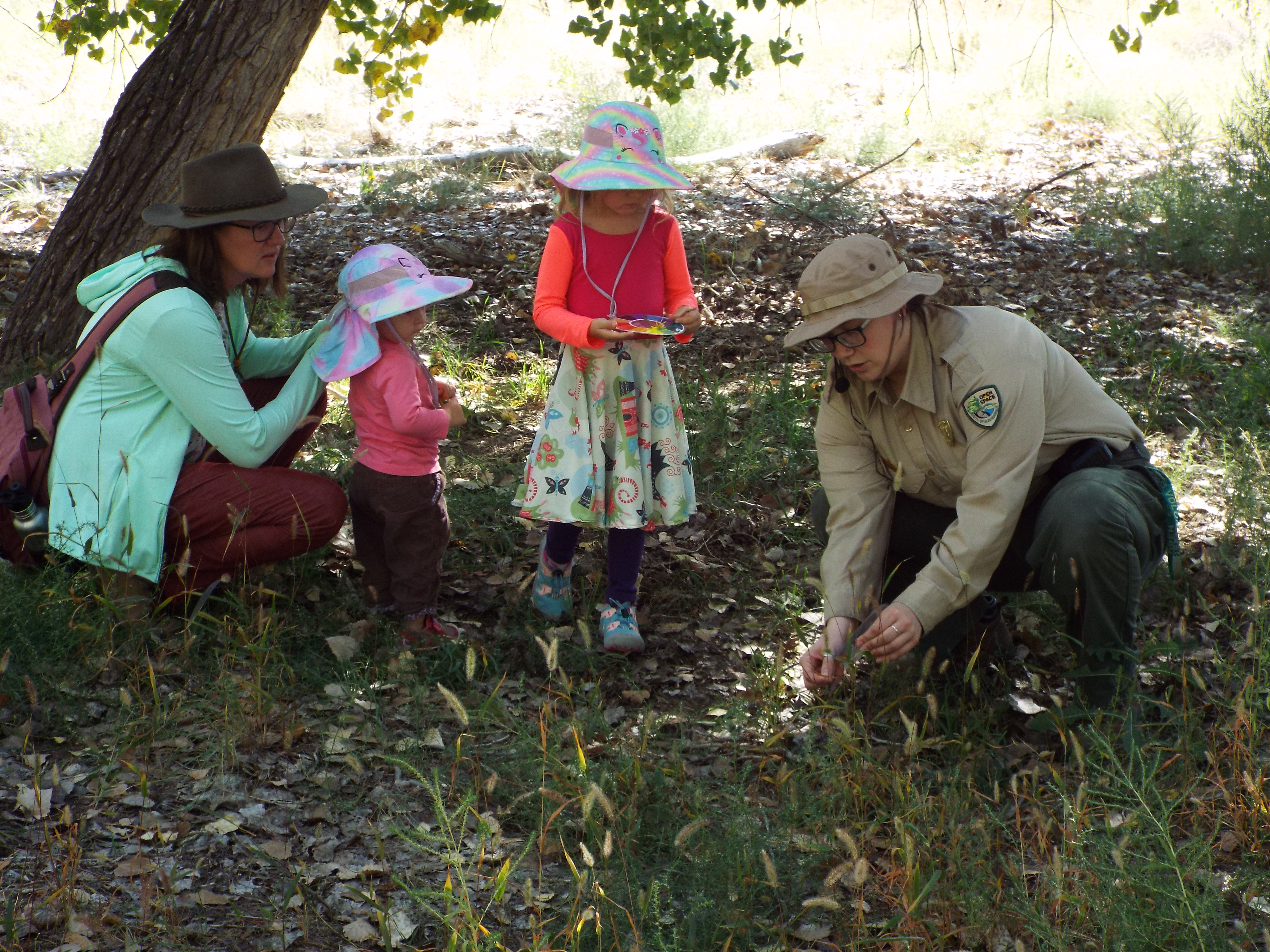 Image of a Bosque trail in broad daylight. Open Space Educator in outdoor uniform kneeling to the right and teaching two youth students in outdoor attire to the left as they observe the vegetation on the ground. Adult supervisor to the left of the students.