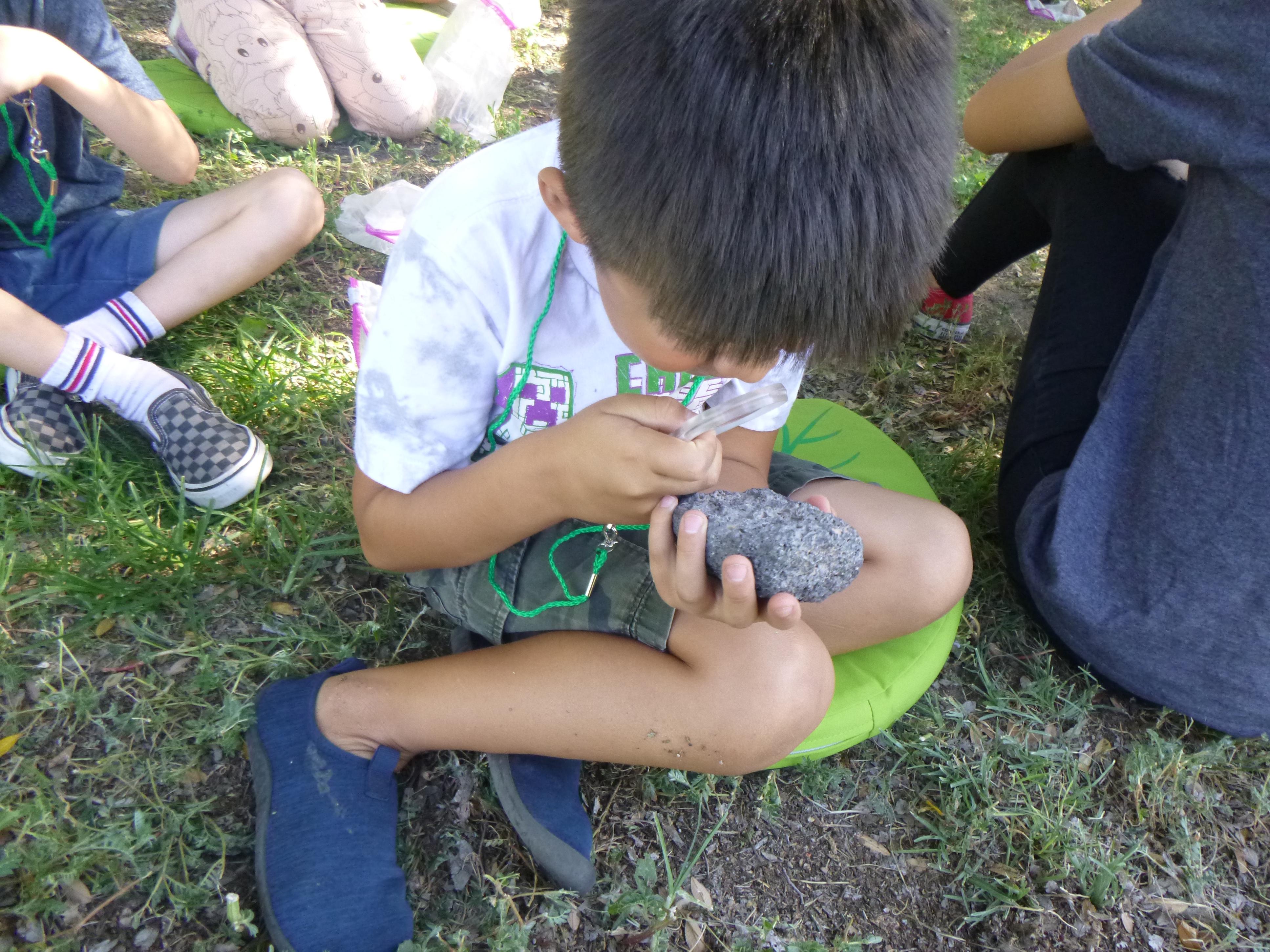 Image of a young student on a patch of grass wearing summer attire, holding a rock on one hand and observing it closely with a magnifying glass on the other.
