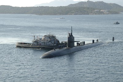 The submarine USS Albuquerque and a tugboat at sea with rocky, shrub-covered hills in the background along with several passing boats in the distance.