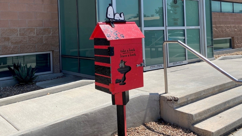 A red doghouse on a pole with Snoopy on top with the words "Take a book leave a book" and a cutout of woodstock the bird on the door.