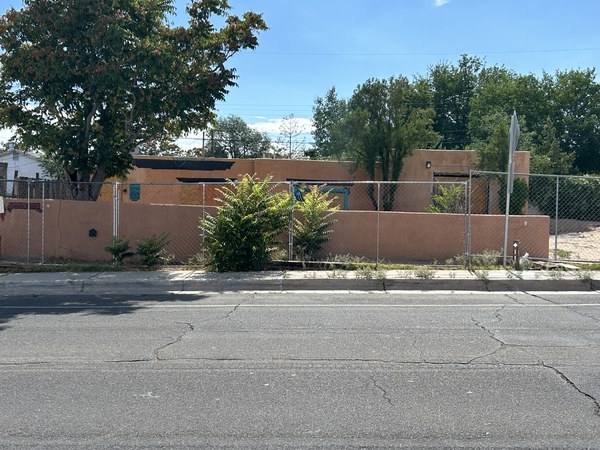 A dilapidated single-story adobe home with an adobe wall, with a chain link fence around the property.