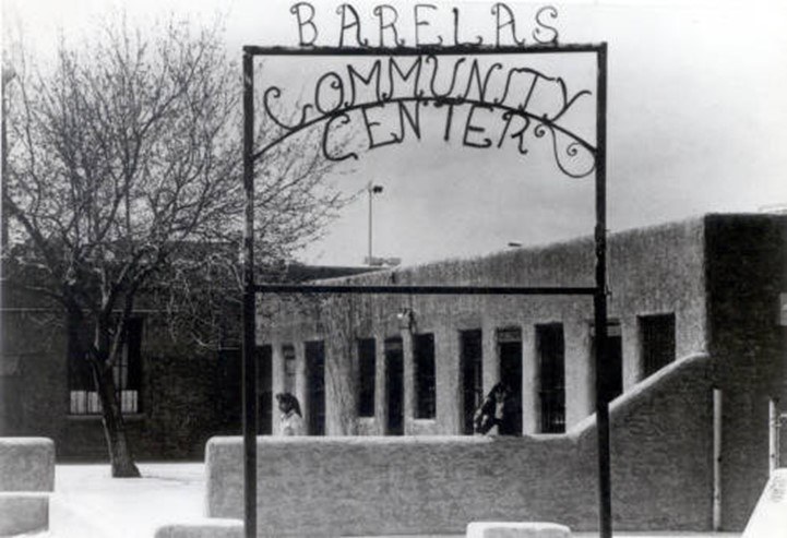A black and white image of a metal sign that reads, "Barelas Community Center" with a traditional adobe building in the background.
