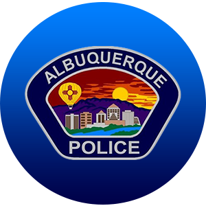 A round graphic with a faded light to blue background and the APD badge logo, which includes the text Albuquerque Police, an illustration of a hot air balloon with the New Mexico state flag, and a sunset behind the Sandia Mountains and the downtown skyline.