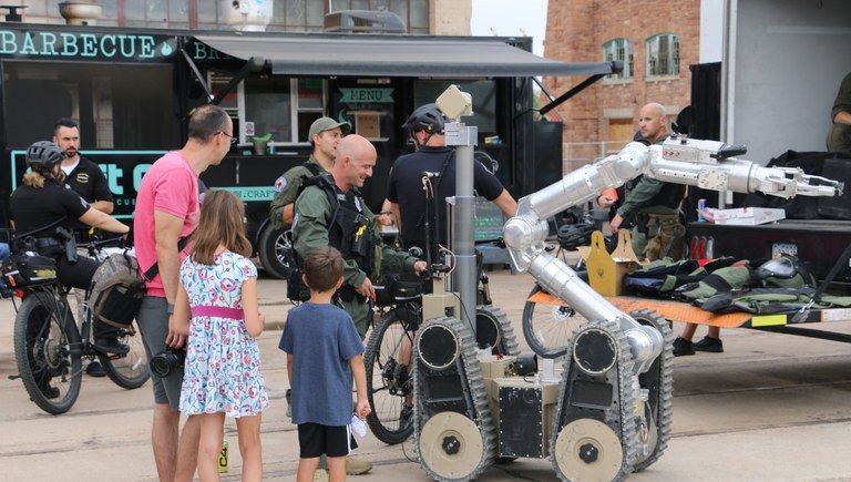 A man and two children look on as a smiling officer operates a robot outside in front of a food truck.