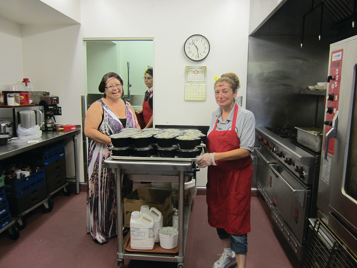 Two women in a Senior Affairs building kitchen pose on either side of a large tray stacked with meals in plastic containers.