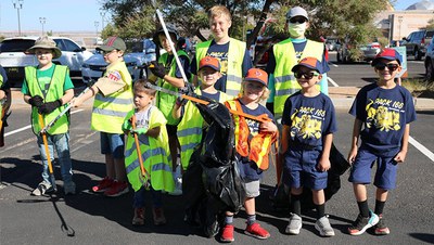 A group of kids, ranging in age from about four to early teens, stands in two rows and smiles at the audience. Most have yellow safety vests on and most are holding trash grabbers and trash bags. Behind them is a parking lot full of cars and a small section of the Balloon Museum can be seen in the upper right.