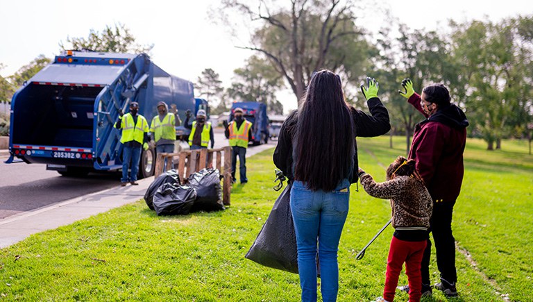 Two adults and a child dressed in hoodies and jeans and holding a trash bag waves at a crew of Solid Waste employees standing in front of a blue Solid Waste truck.