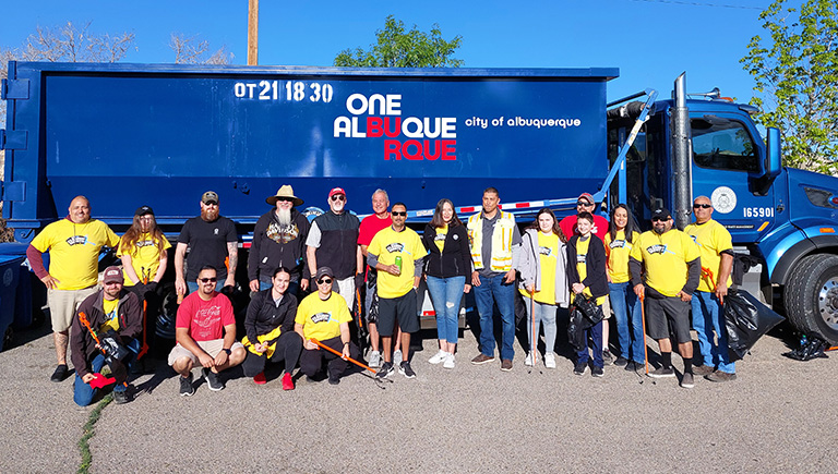 A group of adults stand in two rough rows and smile at the audience. Behind them is a Solid Waste trash truck with the One Albuquerque logo on the side. Bits of trees and a telephone pole can be seen in the background.
