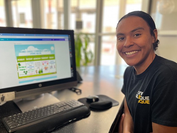 A City of Albuquerque Youth and Family Services employee smiles at the audience. She is seated at a desk with a computer monitor displaying a graphic that is blurred and out of focus.