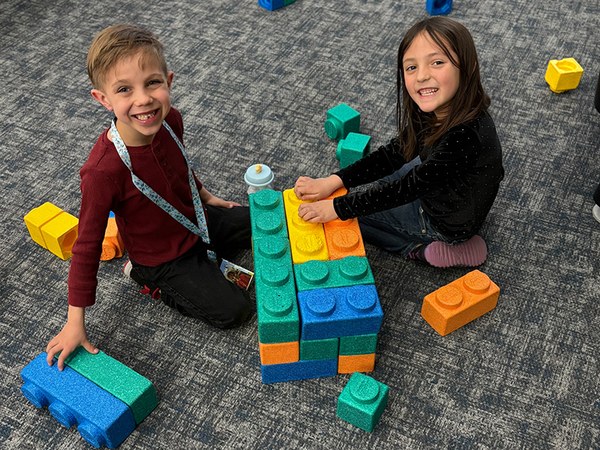 Two children smile up at the audience. They are seated on a gray carpeted floor. In between and around them are large, multi-colored play construction blocks.