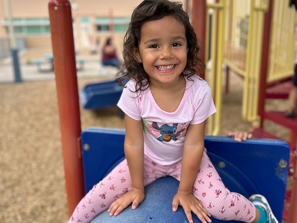 A child poses, smiling at the audience, while sitting on a large blue crawl tube, part of a playground area, with more of the metal and plastic structure visible behind her.