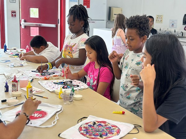 A group of children, gathered on one side of a large table, participate in an arts and craft project creating colorful artworks on white, draw-string bags.