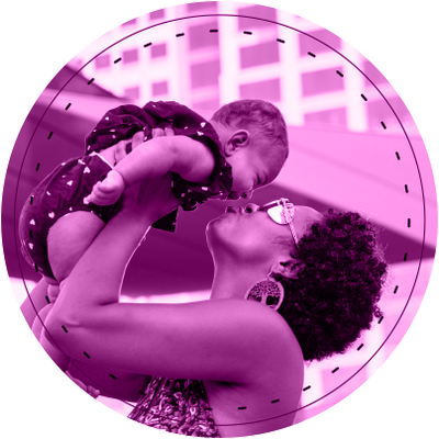 A mother holds up her laughing baby and they embrace nose to nose. The mother has short hair and large earrings with a tree motif. The baby a two piece outfit with a small white heart pattern. In the background are windows of a multi-story building and a section of a sun shade sail.