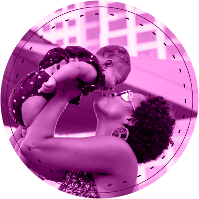 Mother and Child Circle Graphic A mother holds up her laughing baby and they embrace nose to nose. The mother has short hair and large earrings with a tree motif. The baby a two piece outfit with a small white heart pattern. In the background are windows of a multi-story building and a section of a sun shade sail.