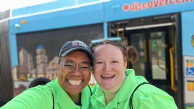 Two people wearing matching bright green shirts pose for a selfie in front of a blue bus with an open door and cityscape artwork.